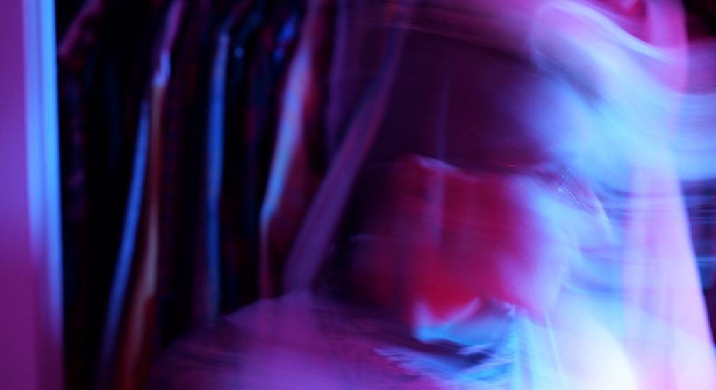 long exposure of Tracy, blurred, looking down and to the right — there are magenta and blue lights all around